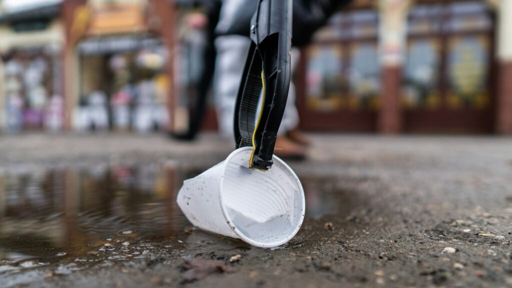Vrijwilliger raapt plastic beker op van natte straat tijdens opruimactie; stijgende opruimkosten zichtbaar in inzet.
